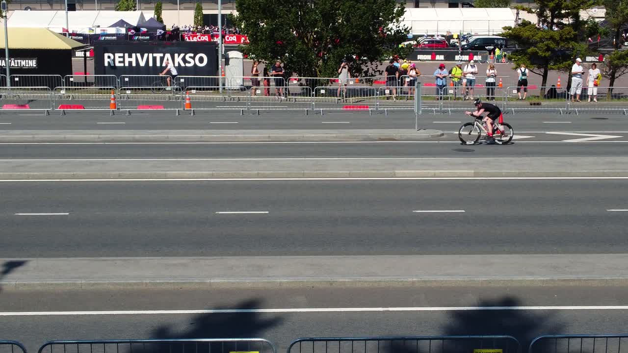 Drone footage captures the tallinn triathlon event with participants on bikes and spectators watching from the sidelines on a sunny day