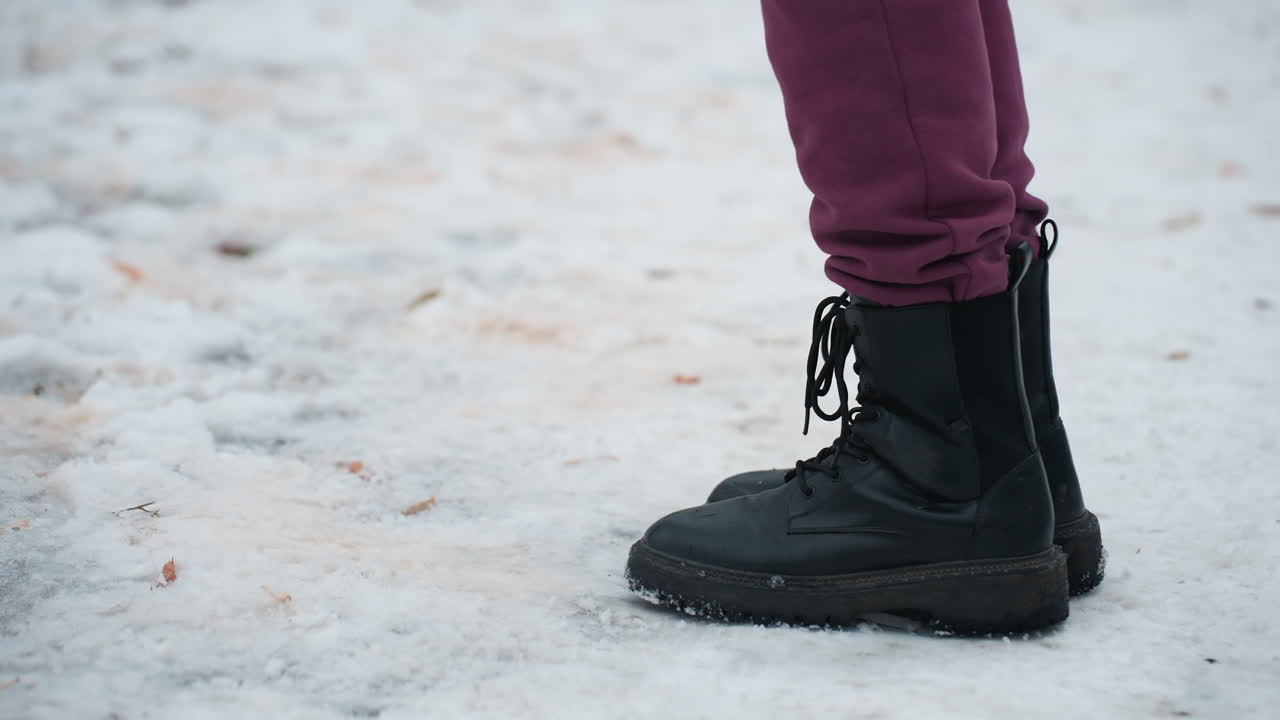 vista lateral de un individuo con botas negras saltando de derecha a izquierda en un terreno cubierto de nieve con follaje seco esparcido, la escena de invierno destaca el movimiento, el calzado y la textura del terreno helado