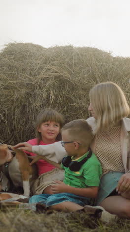 amigos se sientan en la hierba con los niños, cariñosamente acariciando a un perro cerca de la bala de heno en el campo rural, la cálida luz dorada mejora la atmósfera acogedora y alegre, destacando las amistades fuertes, la naturaleza