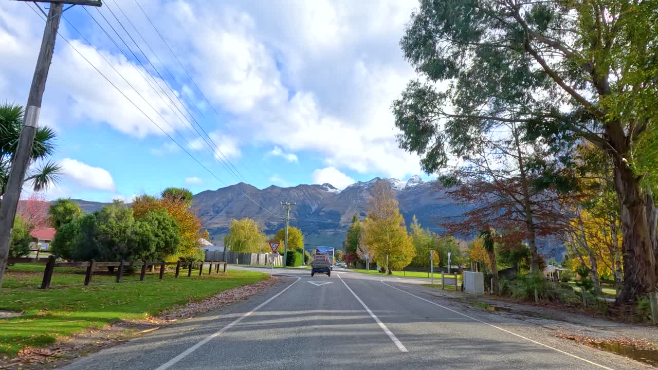 Vehicle drives on rural road past autumn trees, mountains in background, steady forward camera movement