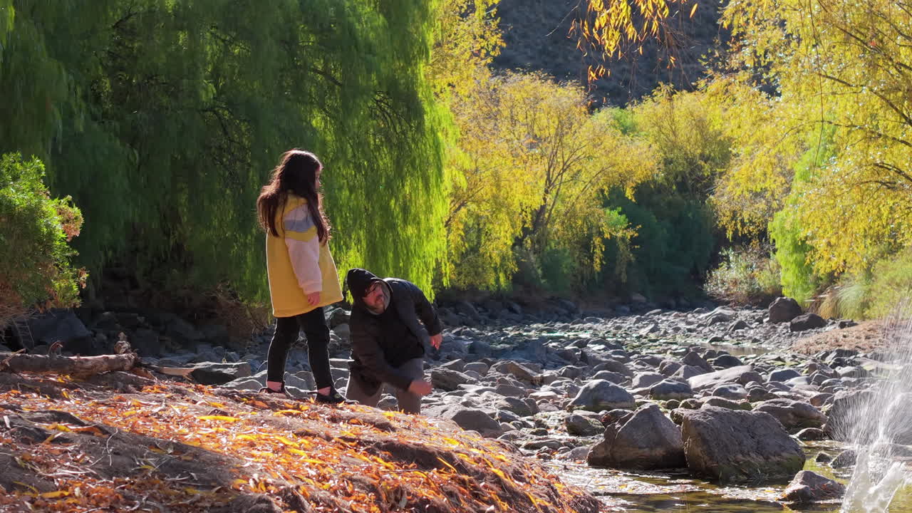 Father and daughter throwing stones in forest riverbed during autumn, family bonding moment