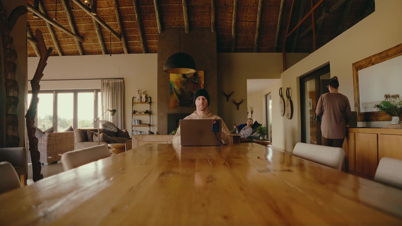 man in beanie sits down at head of large table to work remote on laptop in beautiful wooden home, static 4k
