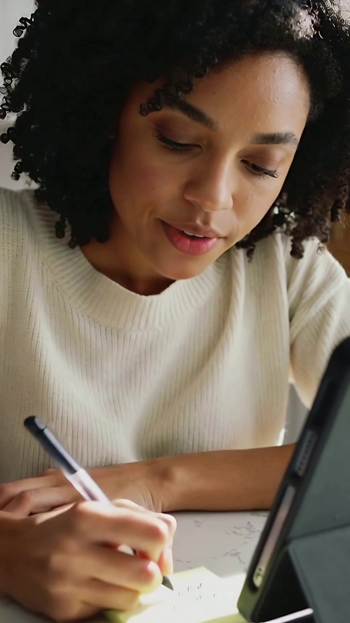 Vertical video: Using tablet, woman in cream top writing yellow notes with black pen in kitchen