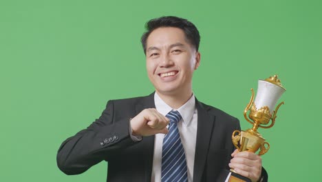 close up of asian business man in a suit and tie with a gold medal showing gold trophy to camera, smiling, and pointing himself being proud being winner on green screen background in the studio