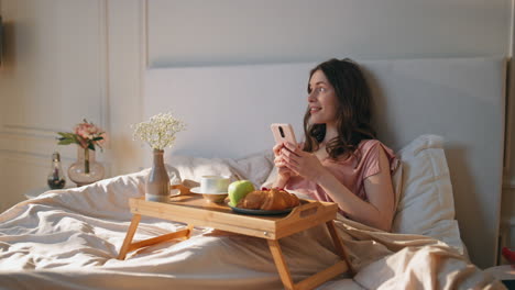 joyful woman browsing mobile phone in comfortable bed. serene girl enjoy morning