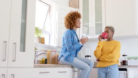 Happy-diverse-female-lesbian-couple-talking-and-drinking-coffee-in-kitchen-in-slow-motion