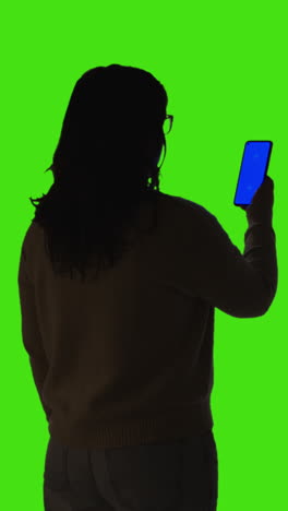 Vertikale-Video-Rückansicht-Einer-Silhouette-Einer-Frau-Mit-Blauem-Mobiltelefon,-Das-Vor-Einem-Grünen-Bildschirm-Steht