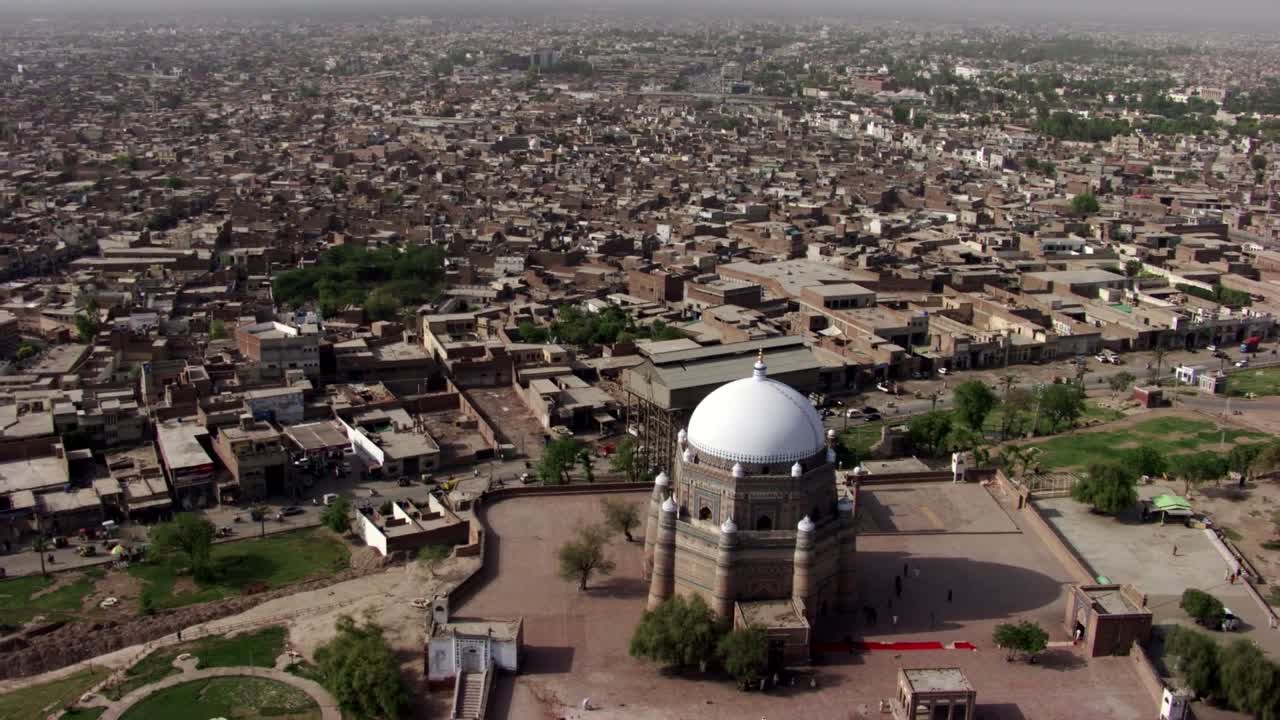 Drone footage captures a aerial view of Multan, Pakistan, Tomb of Shah Rukn-e-Alam in foreground white-domed mausoleum stands out in the dense urban backdrop of residential and commercial buildings