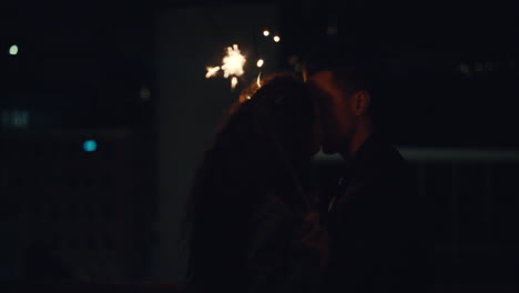 feliz pareja caucásica besándose en la azotea por la noche sosteniendo bengalas celebrando el aniversario disfrutando de la romántica noche urbana de nochevieja
