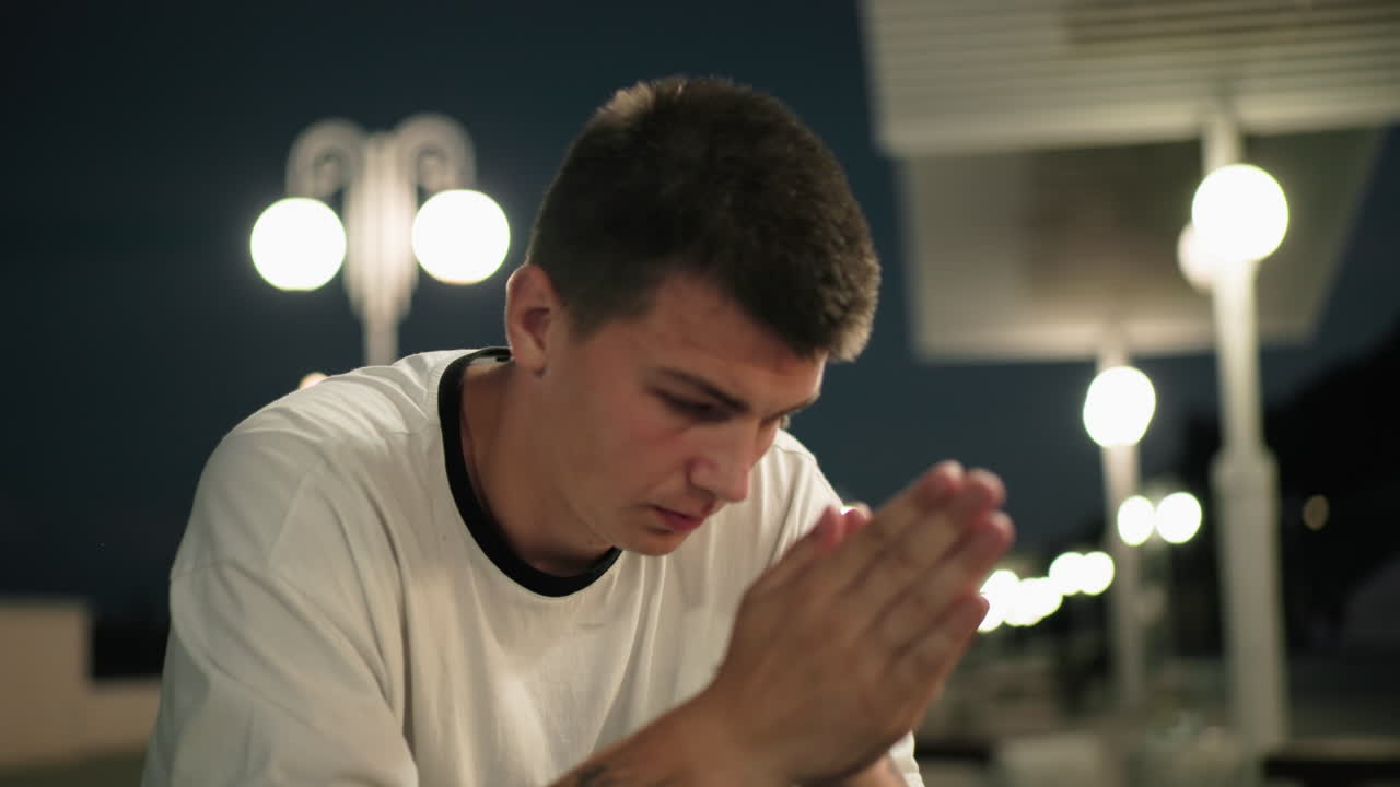 색 폴로를 입은 예술가가 밤에 야외에 앉아서 손으로 얼굴을 고 사려 깊게 문지르고, 거리 램프가 배경에 빛나고 있습니다.