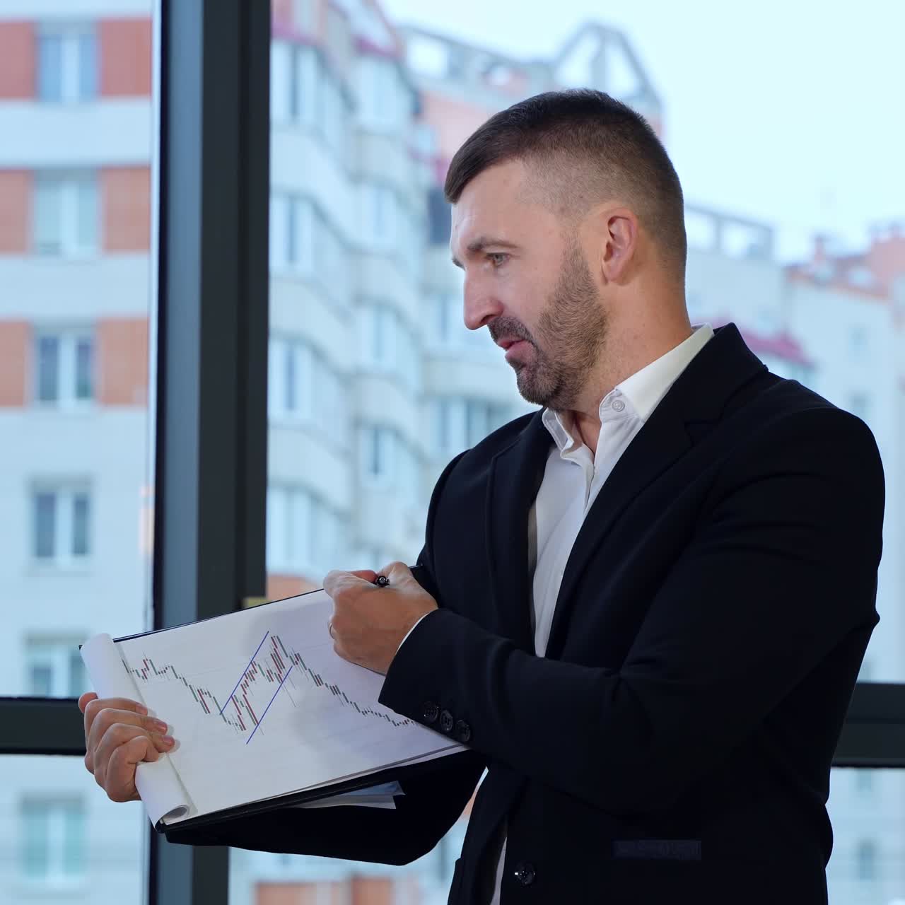 Talkative man shows a graph and comments it to his colleagues. Positive man in official suit explaining charts at office. Big window with house at the backdrop