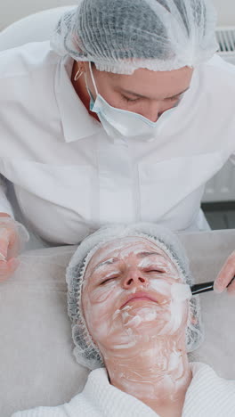 beautician applying peeling moisturizing cream mask on young woman face for rehydrate face skin