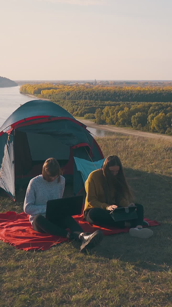 young women with modern tablet and laptop sit near blue tent on hilly river bank against river in windy autumn evening