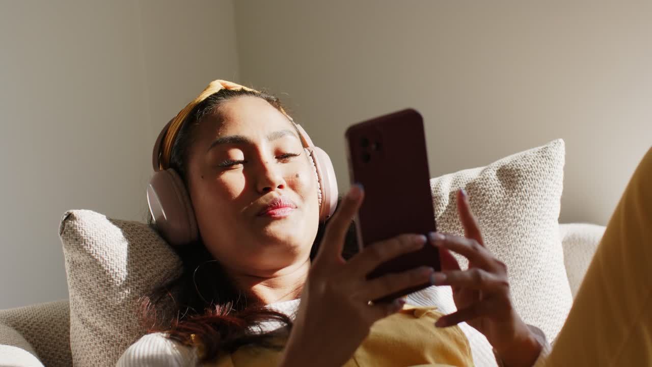 Quando cala il sole, una donna asiatica indossa le cuffie e canta mentre scorre lo smartphone sul divano