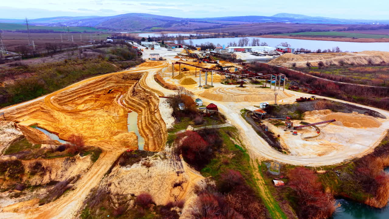 Open pit excavation area with terraced walls, machinery, and water bodies under overcast sky. Aerial panorama of industrial sand quarry and processing site