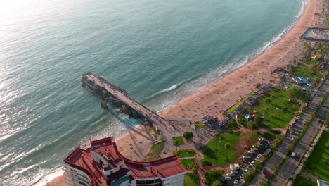 Aerial-Dolly-In-Vergara-Pier,-Türkisfarbenes-Meer-Und-Sandstrand-In-El-Sol-Beach-Zur-Goldenen-Stunde,-Komplexe-Gebäude-Von-Valparaiso,-Chile