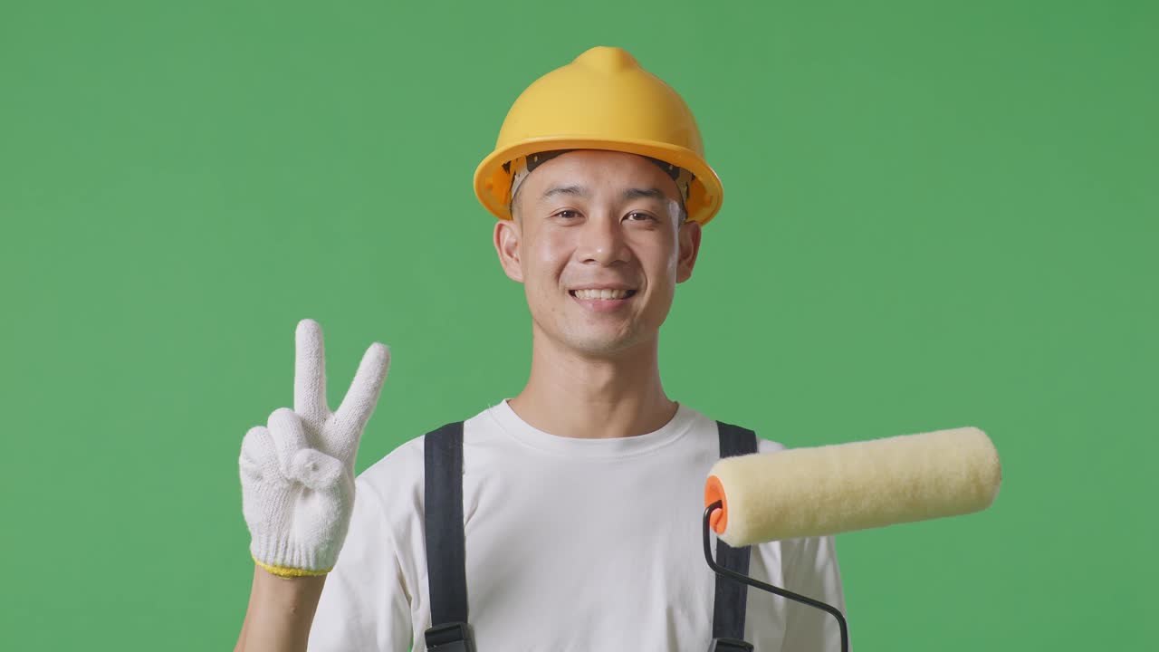 Close Up Of Asian Man Painter Wearing Safety Helmet Smiling And Showing Peace Gesture To Camera While Standing In The Green Screen Background Studio