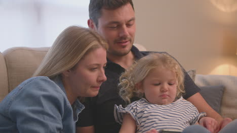 family sitting on sofa at home as young daughter plays game on mobile phone - shot in slow motion