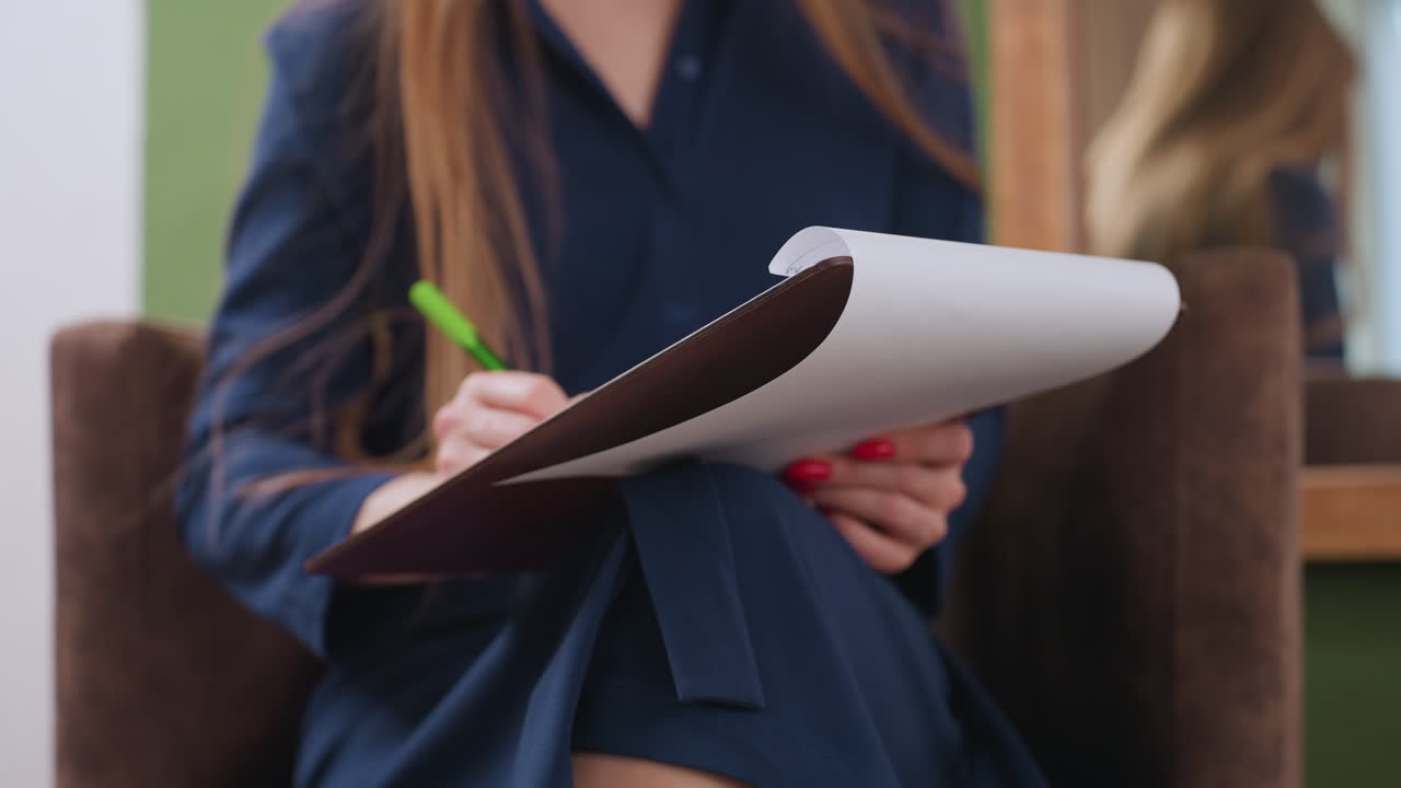 Lower view of emotional coach seated with notepad on lap, writing with pen while engaging in conversation, showing hands, crossed legs, paper, and reflection in mirror in calm indoor setting