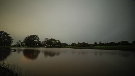 Lapso-De-Tiempo-De-La-Casa-Del-Lago-Con-Luces-Que-Se-Encienden-Y-Apagan-Desde-El-Otro-Lado-Del-Tranquilo-Lago-Reflectante-Por-La-Noche-Con-Nubes-Grises,-Tiro-Estático