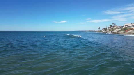 Drone-shot-of-surfers-waiting-for-waves-on-a-sunny-day-with-beautiful-blue-and-green-waves-while-one-surfer-catches-and-rides-a-wave