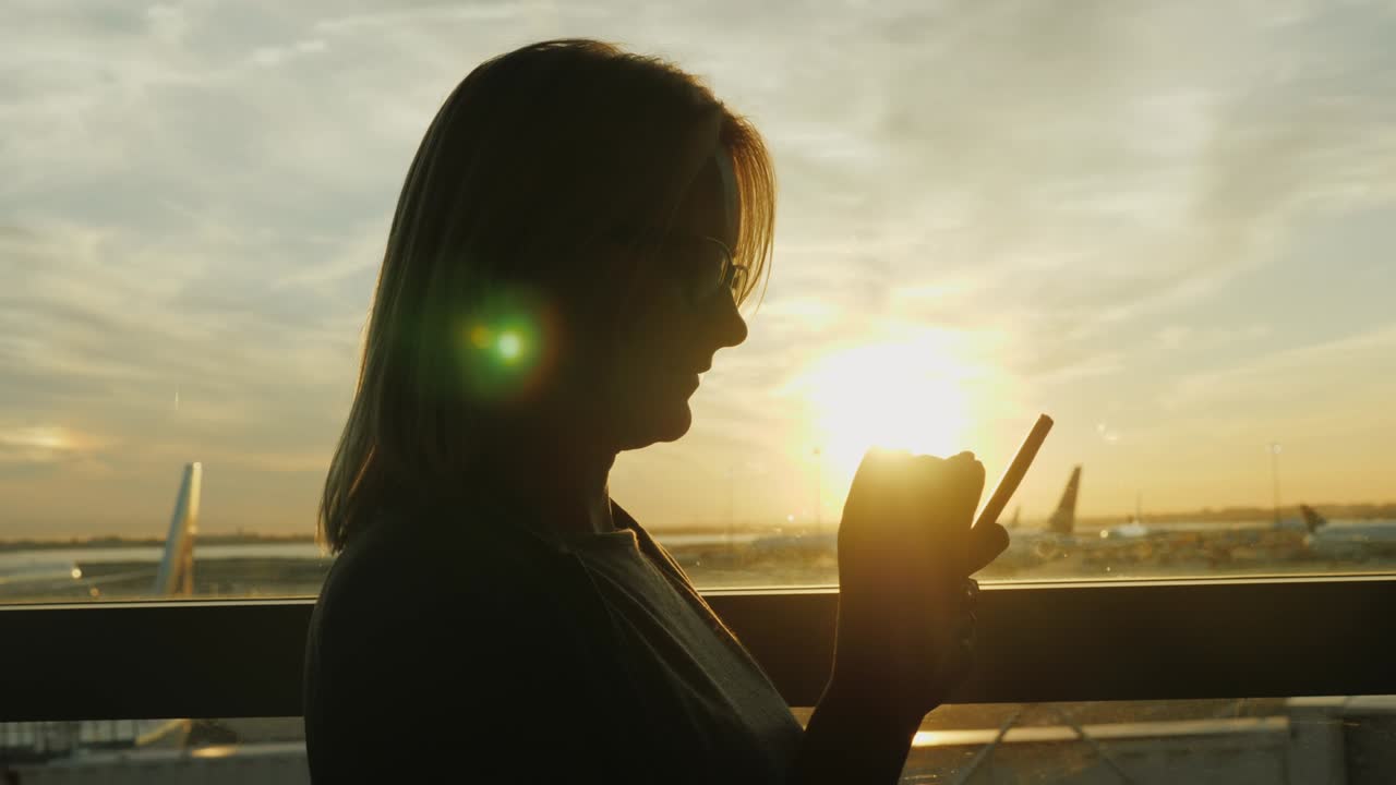 한 여성이 공항의 큰 창문에서 스마트폰을 사용합니다. 해가 지면, 그것을 통해 항공기를 볼 수 있습니다.