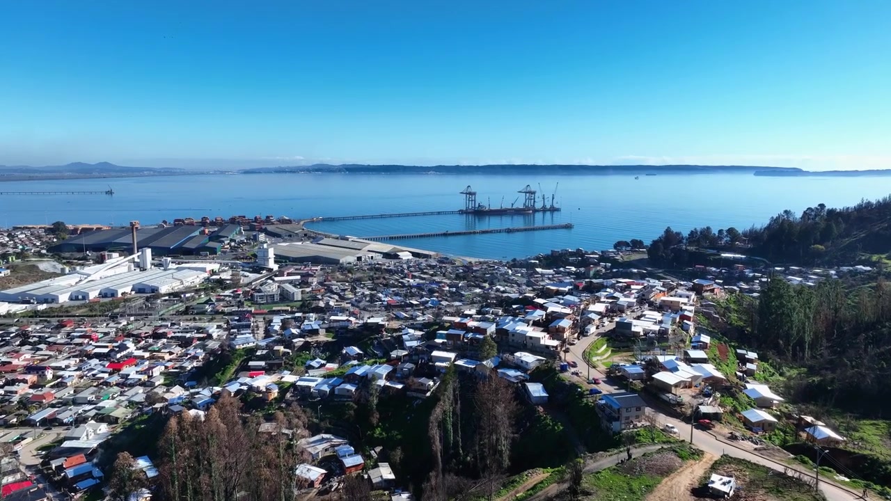 Liitolento tiheän rannikkokaupungin ja teollisuussataman yli, nostureiden ulottuessa pitkin tyyntä lahtea. Lirquén, Penco, Chile.