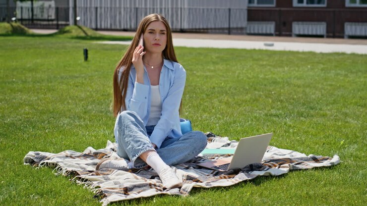 Blid jente som ringer med mobiltelefonen mens hun sitter på campusplenen. Smilende kvinnelig student