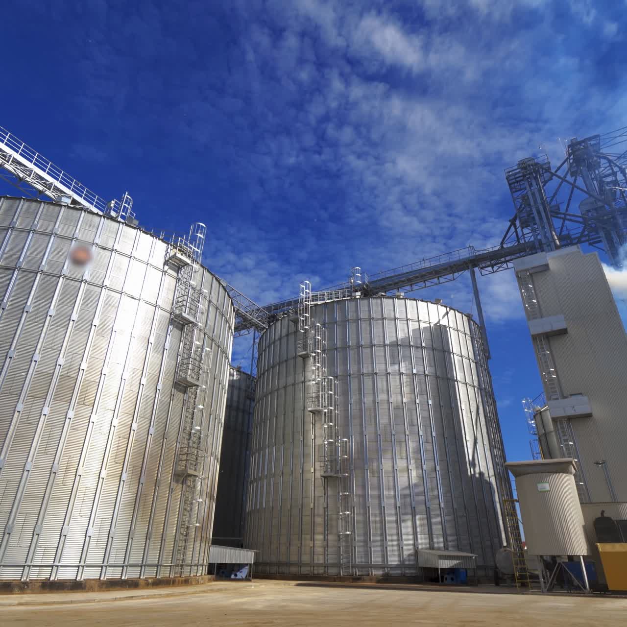 Huge granary outdoors. Large metal elevators on sky background. White smoke released into the air from industrial silos.