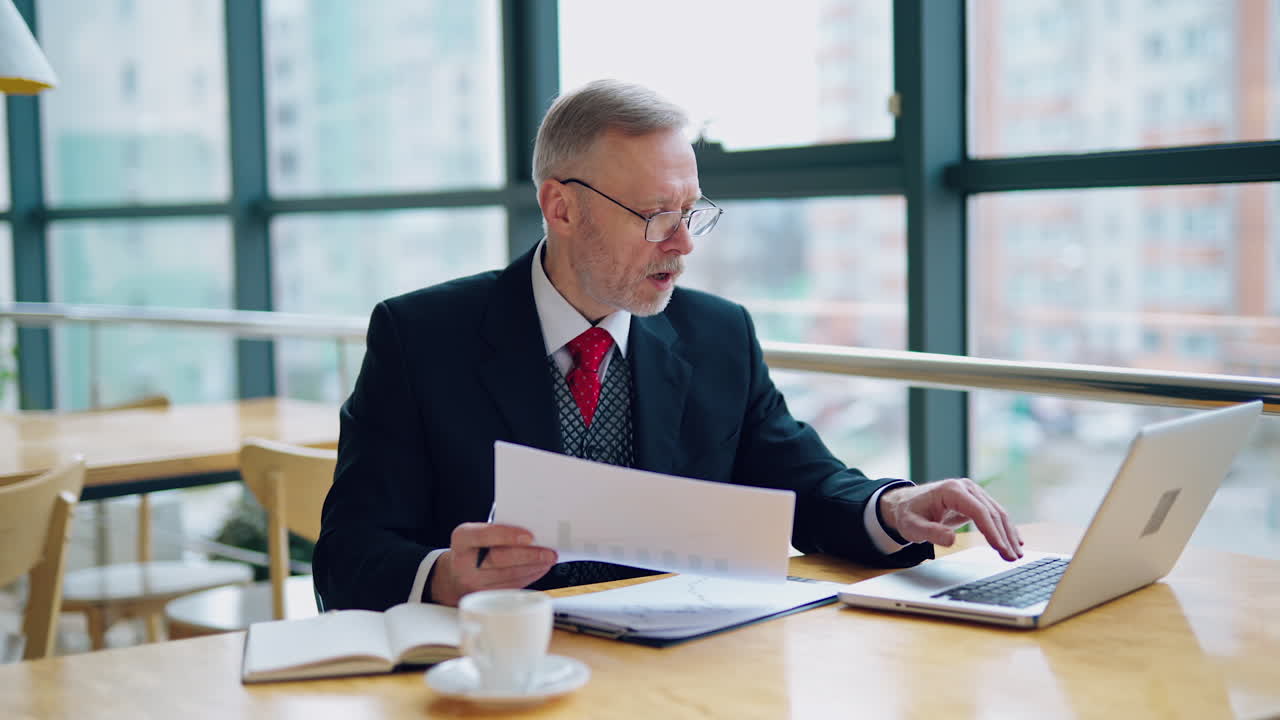 Businessman is checking a printed report or document while sitting at table, working on paper work at a laptop explaining his point of view. Video in an office.