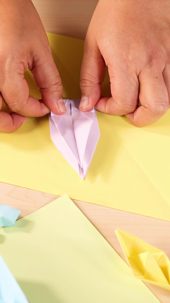 Voksne hænder folder forsigtigt pastel origami-papir, kraftig belysning, nærbillede ovenfra, kreativt arbejdsområde
