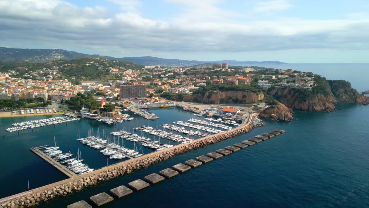 Aerial panorama revealing picturesque sant feliu de guixols nestled along costa brava coastline, showcasing maritime marina, urban architecture, and mediterranean seascape under bright summer sky