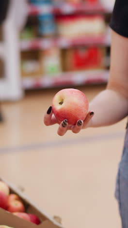 compradora femenina en una tienda de comestibles sosteniendo una bolsa de compras con la mano izquierda, recoge una manzana roja de la exhibición, observándola, alguien que pasa en un fondo borroso en un entorno de tienda vibrante