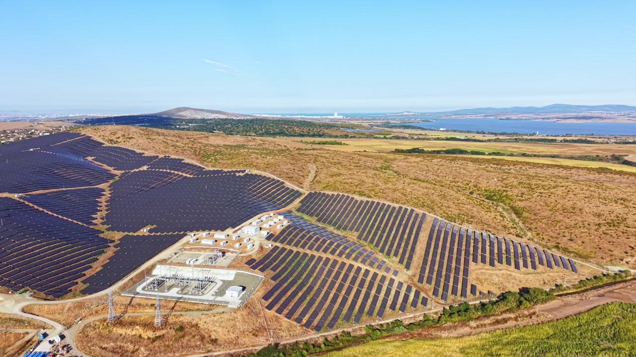 Solcellepark i et landligt landskab med bjerge og vandområde i baggrunden om dagen.