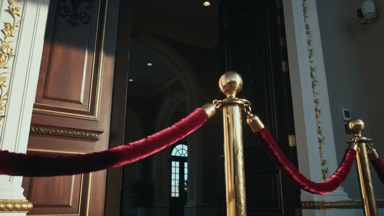 Pulling camera tightening on gold stanchion with red velvet rope at doorway, revealing brass sheen