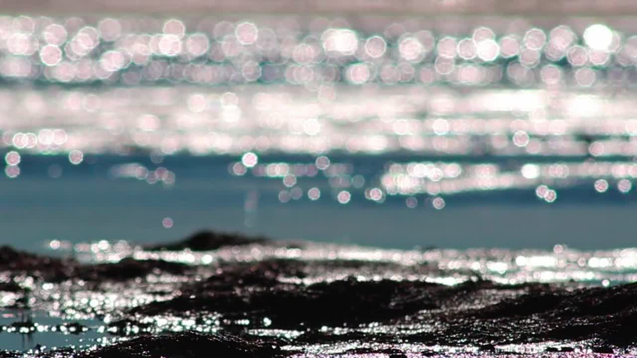 Close-up of shimmering beach water with sparkling reflections and soft bokeh highlights, creating a calm abstract seascape background with bright sunlight glints