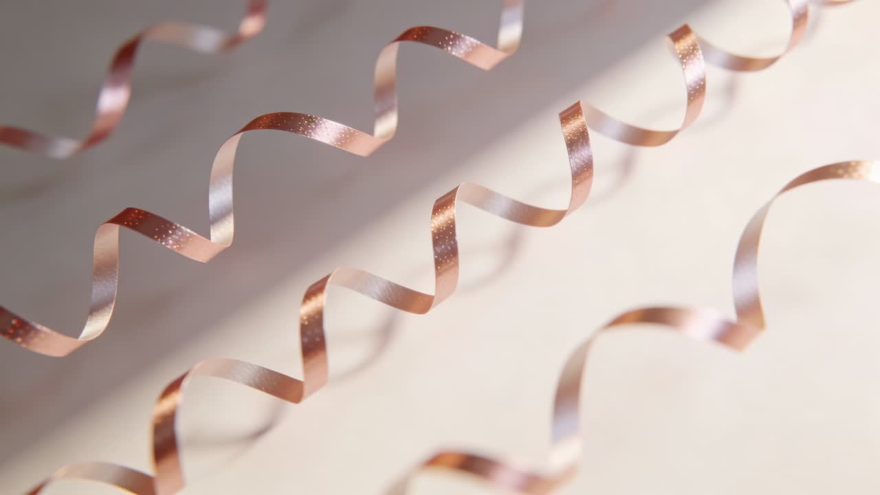 Fitas encaracoladas ouro rosé balançando, projetando sombras curvas em uma superfície fosca pálida, com faixa de luz diagonal