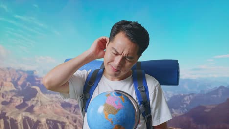 Close Up Of Asian Male Hiker With Mountaineering Backpack Looking At World Globe In His Hands And Making Confused Gesture While Traveling At The Top Of Mountain
