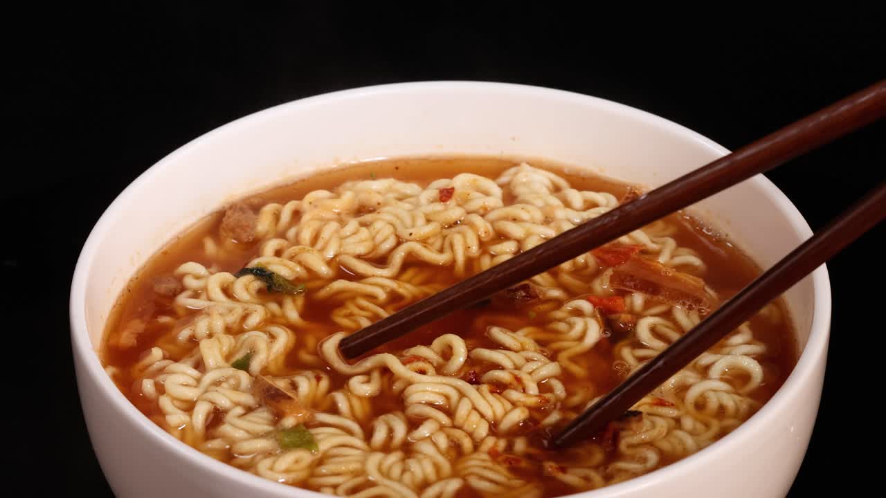 Chopsticks grasp and lift cooked instant noodles from a white bowl filled with broth, under bright studio lighting with a dark, uncluttered background