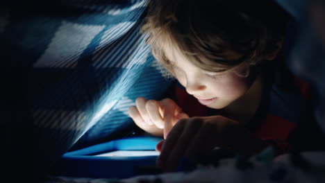 niño feliz usando una tableta digital bajo una manta disfrutando de aprender en la tecnología de pantalla táctil jugando juegos divirtiéndose antes de acostarse