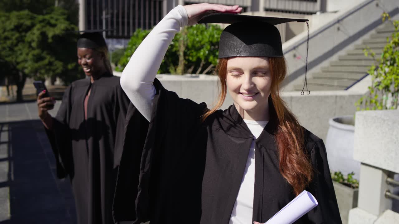 Donne laureate di diverse etnie in toga che si stanno diplomando nel campus, una donna con il diploma che sistema il tocco mentre un'amica scatta foto con il cellulare.