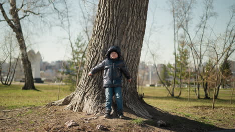 un niño joven se apoya de espaldas contra un gran tronco de árbol, de pie con los brazos extendidos en el árbol, está vestido cálidamente con una chaqueta negra brillante, vaqueros y botas, con árboles vistos a su alrededor