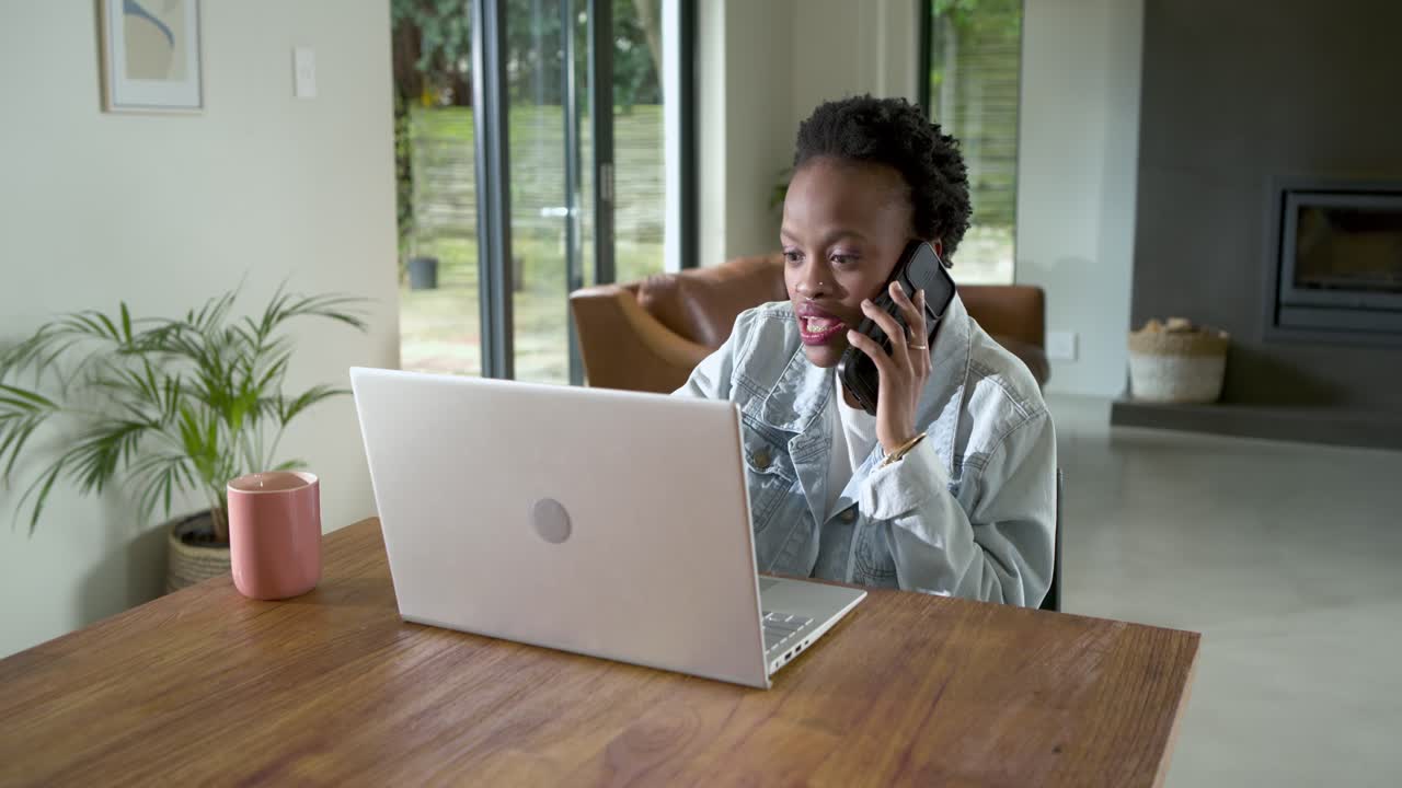 Lifting phone for work call at home office, African American woman typing on laptop between talking