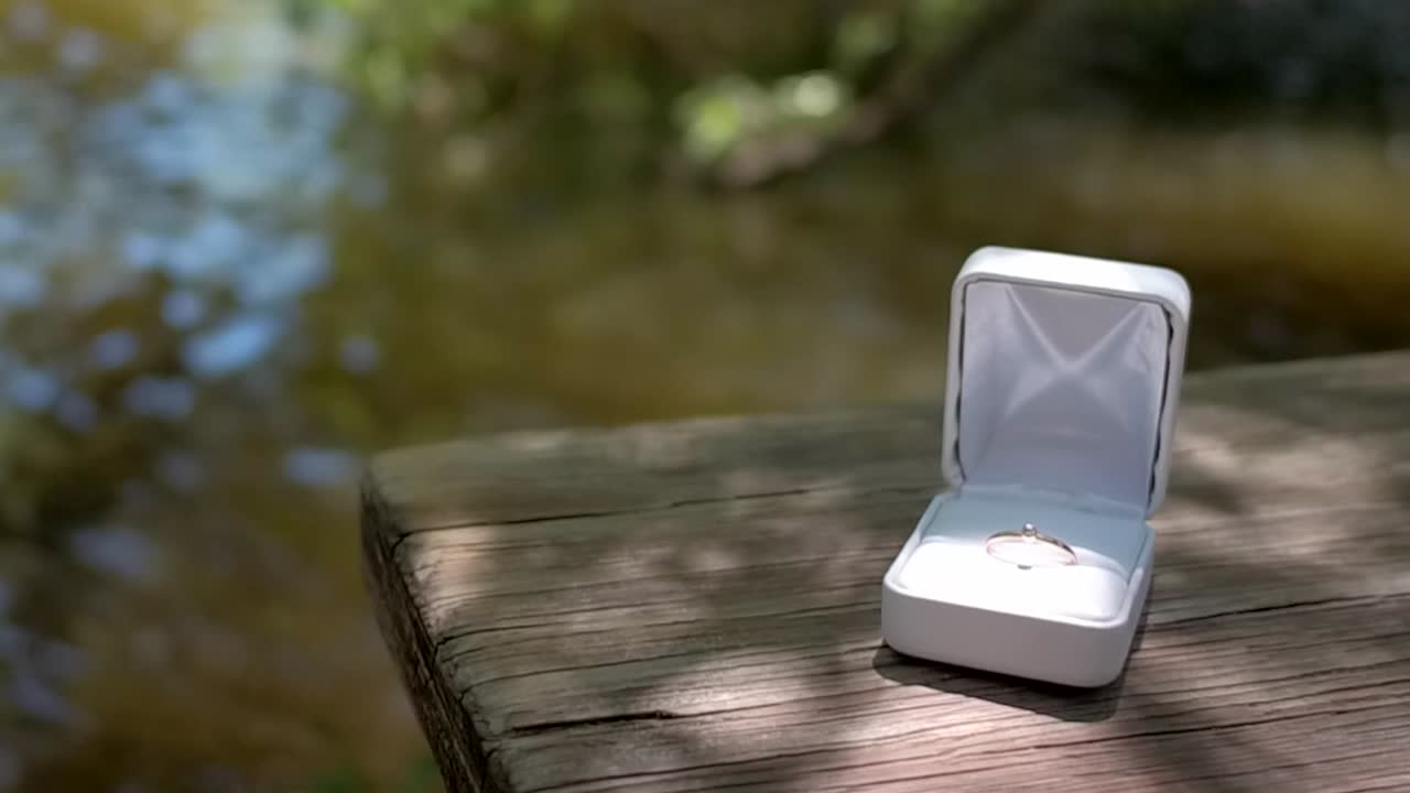Gold engagement ring in white box sits on wooden table by river, dolly push