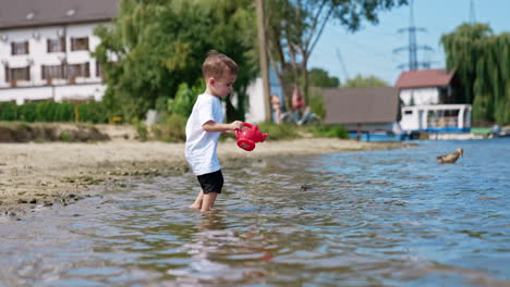 toddler playing with toy watering can and boat in the water by the riverbank