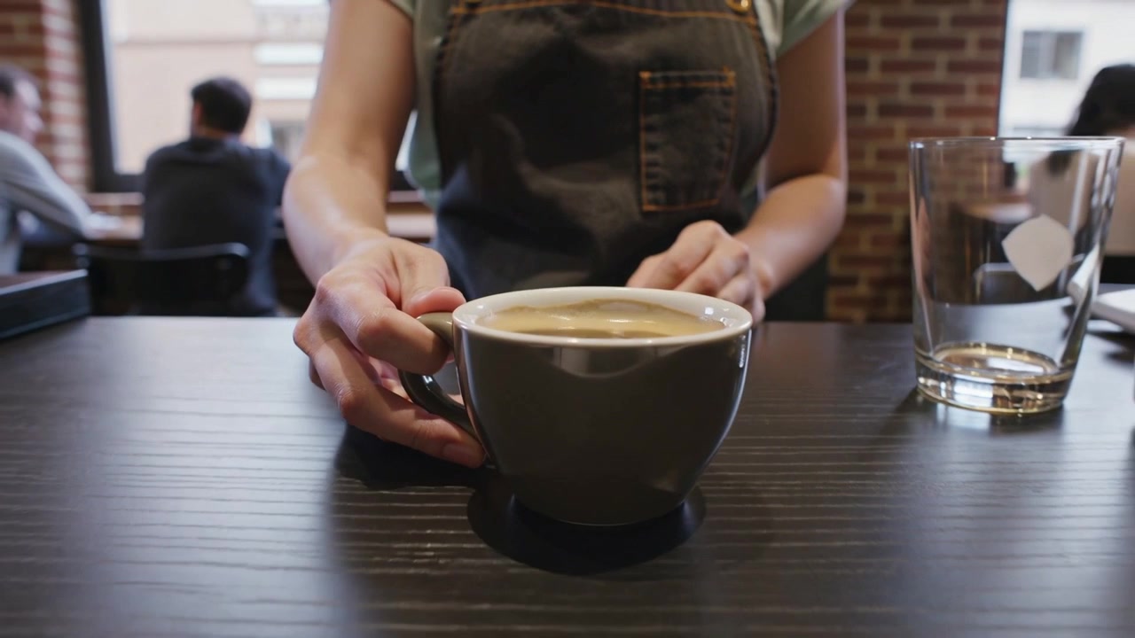 Cameriera con grembiule di jeans che posiziona una tazza di caffè in ceramica sul bancone del bar, su ordinazione del cliente