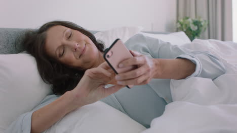 mujer madura feliz usando un teléfono inteligente relajándose en la cama disfrutando de navegar en línea enviando mensajes de texto compartiendo estilo de vida