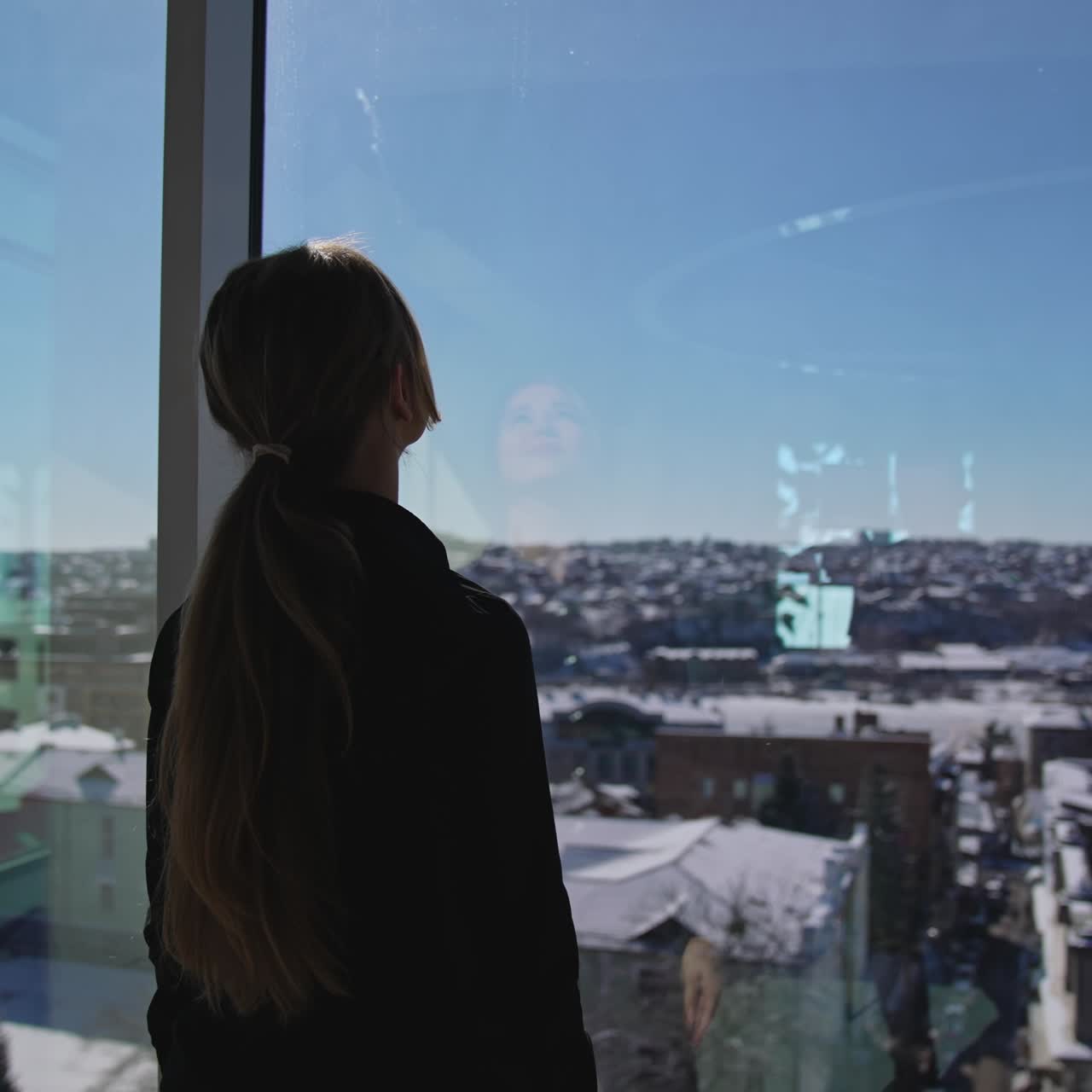 Young good-looking female wearing dark clothes standing in front of the panoramic windows. Sunrays coming into the big office room. Girl looking out of the window and enjoying the cityscape