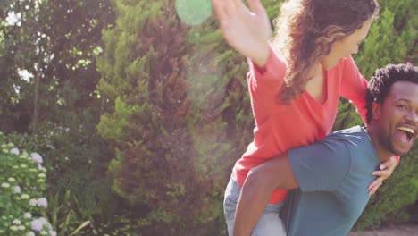 Happy-biracial-man-holding-piggyback-his-fiance-with-engagement-ring-on-hand-in-garden-in-sun