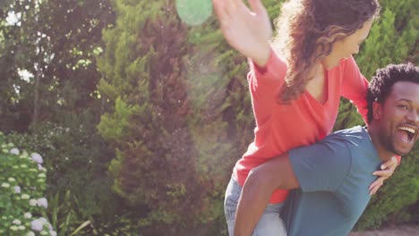 feliz hombre biracial sosteniendo a su prometida con anillo de compromiso en la mano en el jardín al sol
